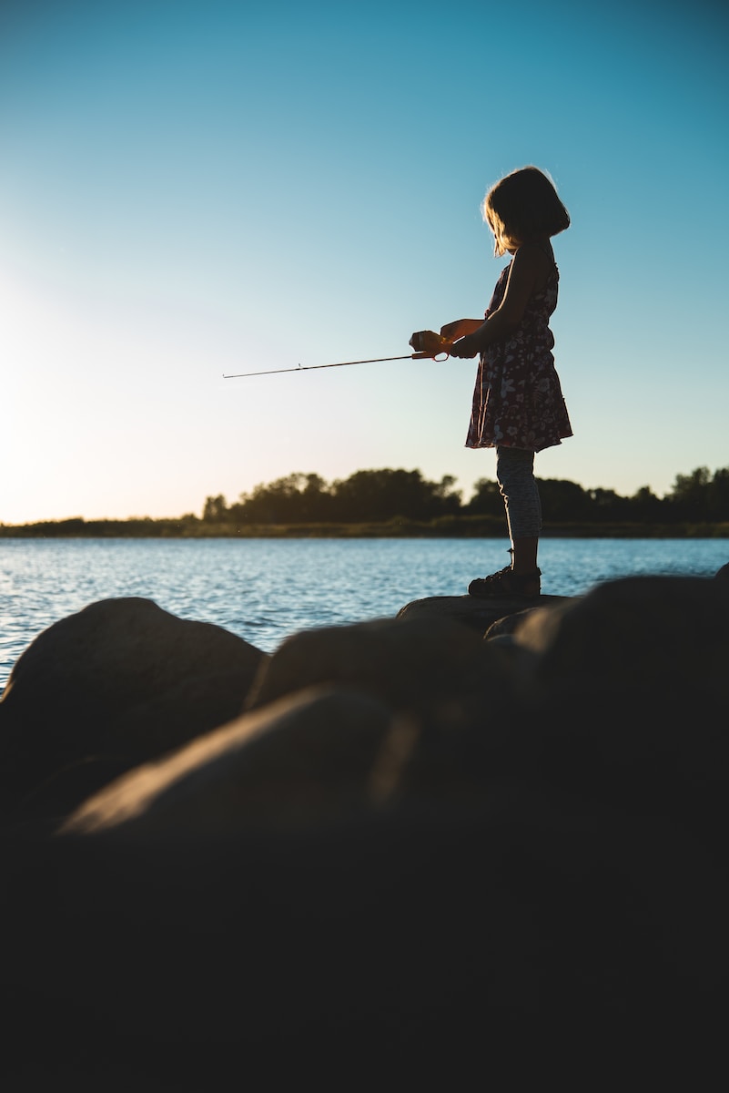 woman in white and black dress holding fishing rod standing on rock near body of water